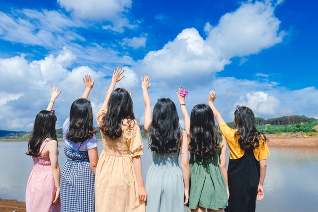 six girls facing the waters while raising their right hands like reaching for the sky